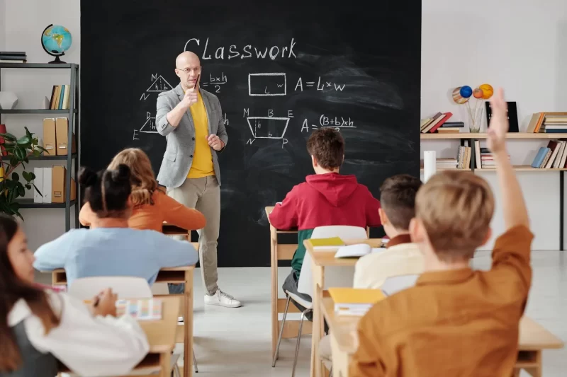 Teacher giving classes at classroom with children, kid raising hand.